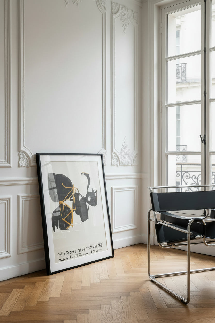 Felix Droese, Galerie Rudolf Zwirner exhibition poster (1982), shown framed, leaning against a white panelled wall beside a tubular steel chair.